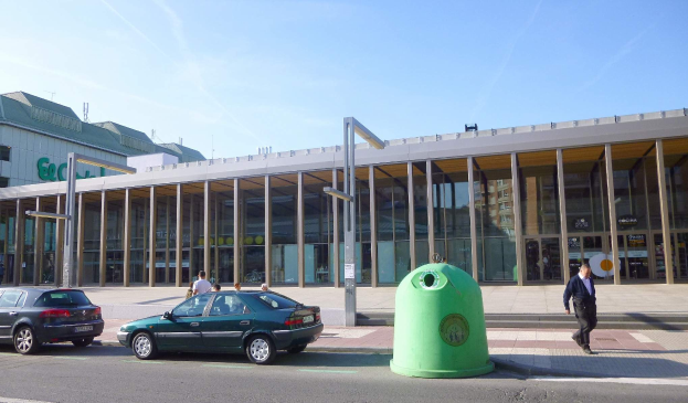 Grüner Müllcontainer auf einer Stadtstraße neben einem gläsernen Gebäude mit Säulen, mit Fahrzeugen und Passanten in der Nähe, unter einem bewölkten Himmel am Berliner Hauptbahnhof, der nun ein umweltfreundliches Einkaufszentrum ist.
