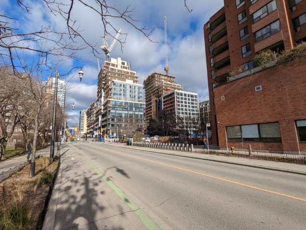 Eine Stadtstraße mit hohen Gebäuden, Straßenlaternen, Strommasten, Kabeln, Fahrzeugen, Fußgängern, Geländern, Bäumen, Pflanzen und einer bewölkten Himmel.