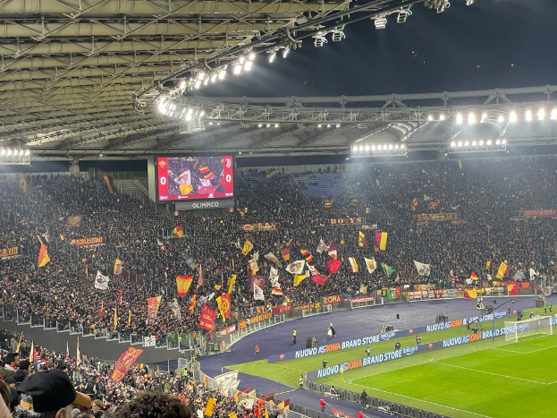 Eine große Menge in einem hell erleuchteten Stadion schwenkt Fahnen und Banner, wobei einige Besucher Mützen tragen, während sie auf ein Fußballspiel warten.