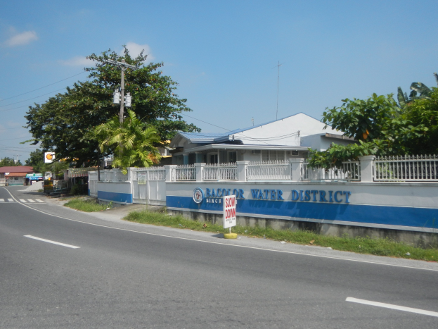 Eine Straße mit einem "Wasserstadt"-Schild, Gras, einem Zaun, einer Wand mit Text, Bäumen, Häusern mit Dächern und Fenstern, Masten mit Drähten und einem bewölkten Himmel.