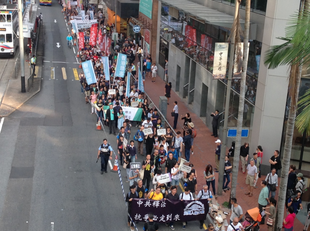 Eine große Gruppe von Menschen marschiert auf einer Straße während einer Demonstration in Hong Kong, mit Schildern und Plakaten.