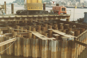 Baustelle mit einem Kran, der Stahlstangen von einem großen Haufen in der Nþhe von Wasser anhebt, umgeben von Gebüuden, Büschen und einem klaren blauen Himmel.