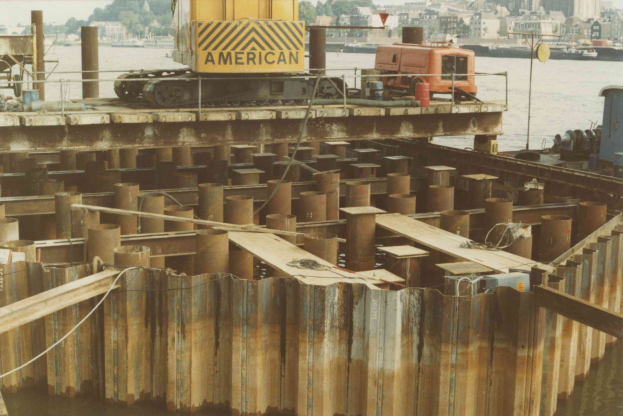 Baustelle mit einem Kran, der Stahlstangen von einem großen Haufen in der Nþhe von Wasser anhebt, umgeben von Gebüuden, Büschen und einem klaren blauen Himmel.