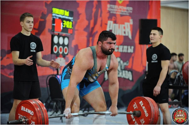 Ein Mann, der eine Hantel in einem Gewichtheben-Wettkampf hochhebt, umgeben von Zuschauern, mit Wettkampfausrüstung und Hinweisschildern, die die 2018 World Weightlifting Championships anzeigen.