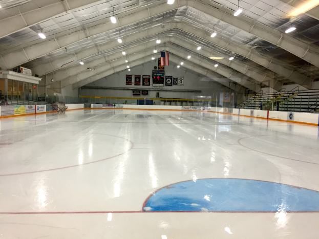 Großes Indoor-Eisstadion mit Deckenbeleuchtung, Werbetafeln, einer Rückwand, Treppen mit Geländern und einigen Menschen, was auf eine Sportanlage hinweist, die likely für Hockey genutzt wird.