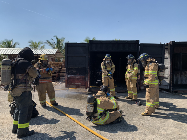 Feuerwehrleute in Helmen und Zylindern stehen vor einem Gebäude und halten eine Leitung in der Hand, mit Bäumen, Schuppen und hölzernen Gegenständen im Hintergrund, während einer Feuerübung.