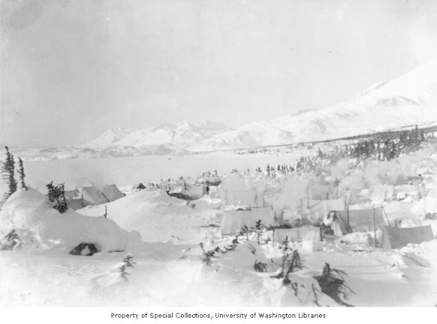 Ein Schwarz-Weiß-Foto einer Gruppe von Zelten im Schnee, umgeben von Bäumen und Bergen, mit dem Text "Kootenay Camp, Alaska, circa 1900" unten.