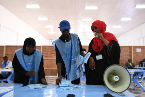 Eine Gruppe von Menschen steht um einen Tisch mit einem Megafon, Papieren und anderen Gegenständen, während andere im Hintergrund auf Stühlen oder stehen, unter einer Decke mit Lampen, was auf eine Wahllokalität hindeutet.