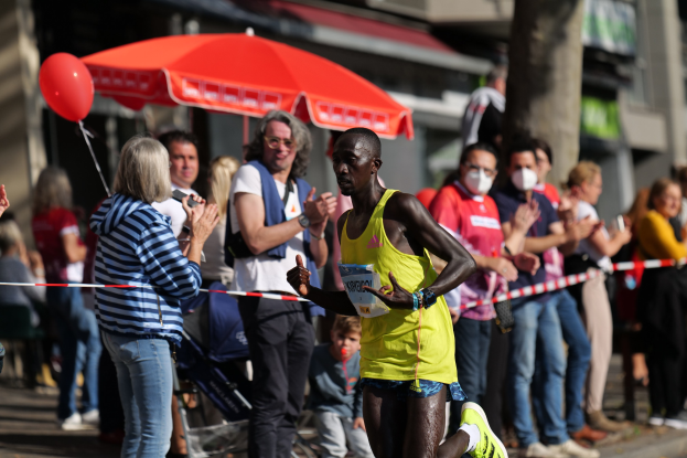 Ein Mann in einem gelben T-Shirt und Laufschuhen sprintet durch einen Marathon, während ihn eine Menge umgibt, mit einem Schirm und einem Kinderwagen im Hintergrund.