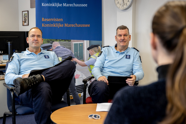 Zwei Polizeibeamte sitzen an einem Tisch mit einer Frau, umgeben von Dokumenten und Gegenständen, mit einem wandmontierten Banner, einer Uhr und Fotorahmen im Hintergrund und einem weiteren Tisch mit einem Monitor und Zubehör links daneben.
