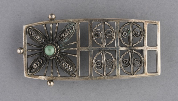 Ein silberner Gürtelschnalle mit einem grünen Stein in der Mitte auf einer aschfarbenen Oberfläche.