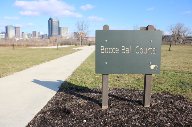 Ein Schild am Boden in der Nähe eines Weges und Bäume trägt die Aufschrift "Bocce Ball Courts in Indianapolis, Indiana" mit einem bewölkten Himmel darüber.