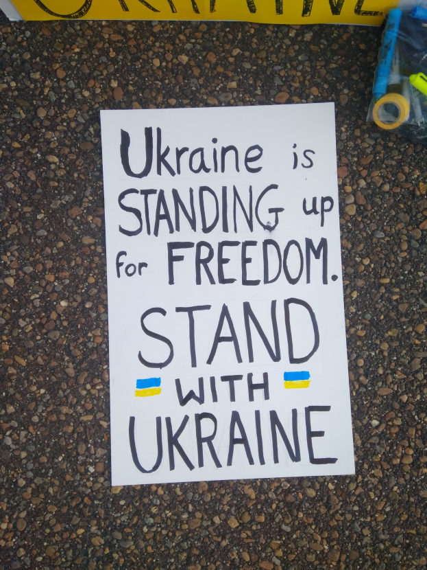 Ein weißer Zettel mit der Aufschrift "Ukraine steht für Freiheit auf, stell dich Ukraine" liegt vor einer gelben Fahne auf dem Boden, rechts daneben eine Plastikabdeckung mit verschiedenen Gegenständen.