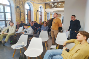 Eine Gruppe von Menschen sitzt in einem Raum mit einer Wand, Fenstern und Deckenlampen, einige halten Gegenstände, während sie über die Zukunft des Designs in Berlin diskutieren.