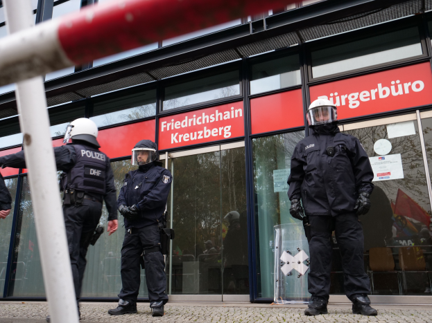 Eine Gruppe von Polizisten in Helmen und Handschuhen steht vor einem gläsernen Gebäude, mit einem Pfahl links und Schildern im Hintergrund, die "Friedrichshain Kreuzberg" und Papiere an den Glaswänden angeklebt zeigen.