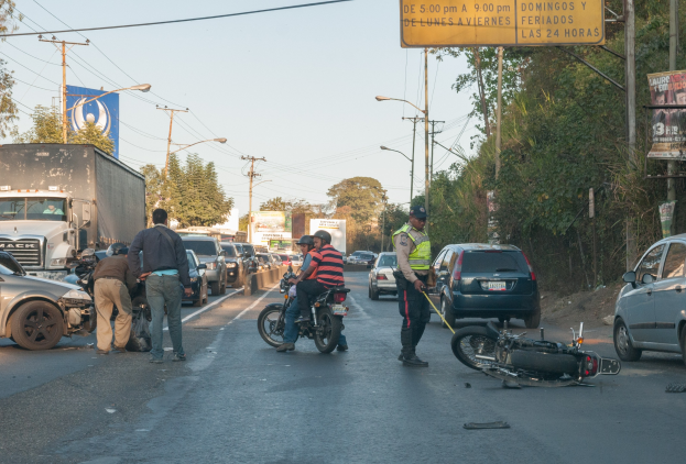 Eine Gruppe von Menschen steht um ein verungl"ucktes Motorrad auf der Seite einer Stra{ße mit vorbeifahrenden Fahrzeugen, B"umen, Strommasten und einem klaren Himmel im Hintergrund.