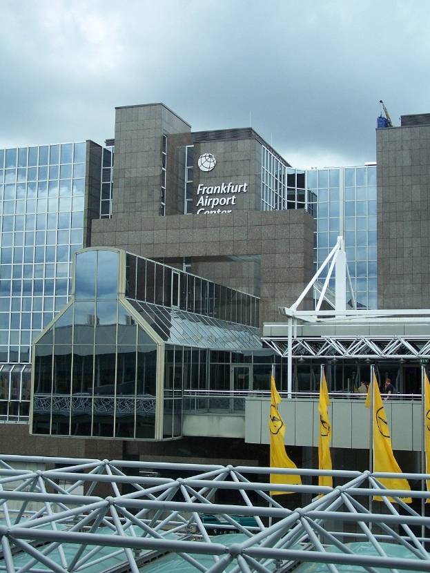 Frankfurt Airport in Frankfurt, Deutschland, mit einem großen Glaswandgebäude mit Text, umgeben von gelben Flaggen und Eisenstangen unter einem bewölkten Himmel.