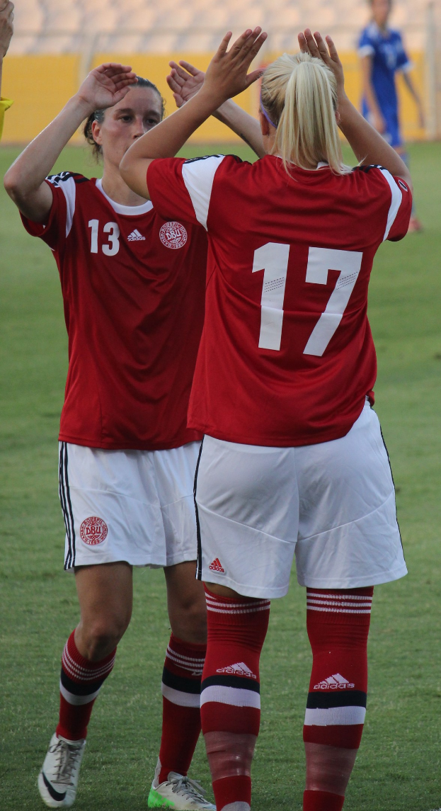 Zwei Frauen in roten Hemden und weißen Shorts klatschen auf einem Fußballfeld mit einer Person in einem blauen Kleid, einer gelben Tafel und Stühlen im Hintergrund.