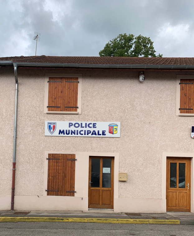 Ein Polizeigebäude der Stadt mit einem Schild, Fenstern, Türen, einer Namensplakette, einem Rohr, einem Baum und einem bewölkten Himmel im Hintergrund.