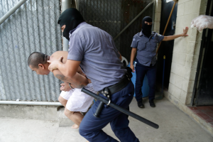 Ein Mann wird von zwei Polizisten vor einem Gebäude eskortiert, wobei einer der Beamten den Mann festhält und der andere einen Stock hält, mit einer Wand, einer Tür, einem Rohr und einem Netz im Hintergrund.