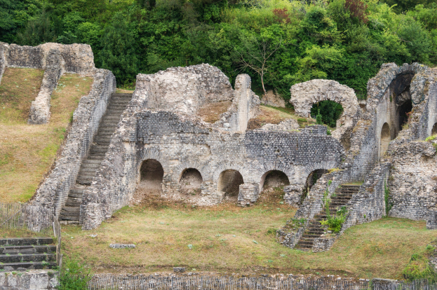 Römerzeitliche Amphitheaterruinen mit Treppen, Mauern und Bögen auf einer grünen Wiese, umgeben von einem Zaun mit Bäumen im Hintergrund und einem Wasserzeichen auf dem Bild.