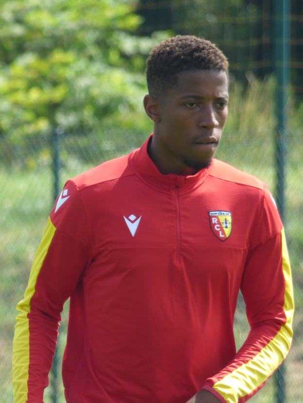 Ein junger Mann in rot-gelbem Trikot steht auf einem Fußballfeld, umgeben von einem Zaun und Bäumen, mit entschlossener Miene, wahrscheinlich ein Flügelspieler.