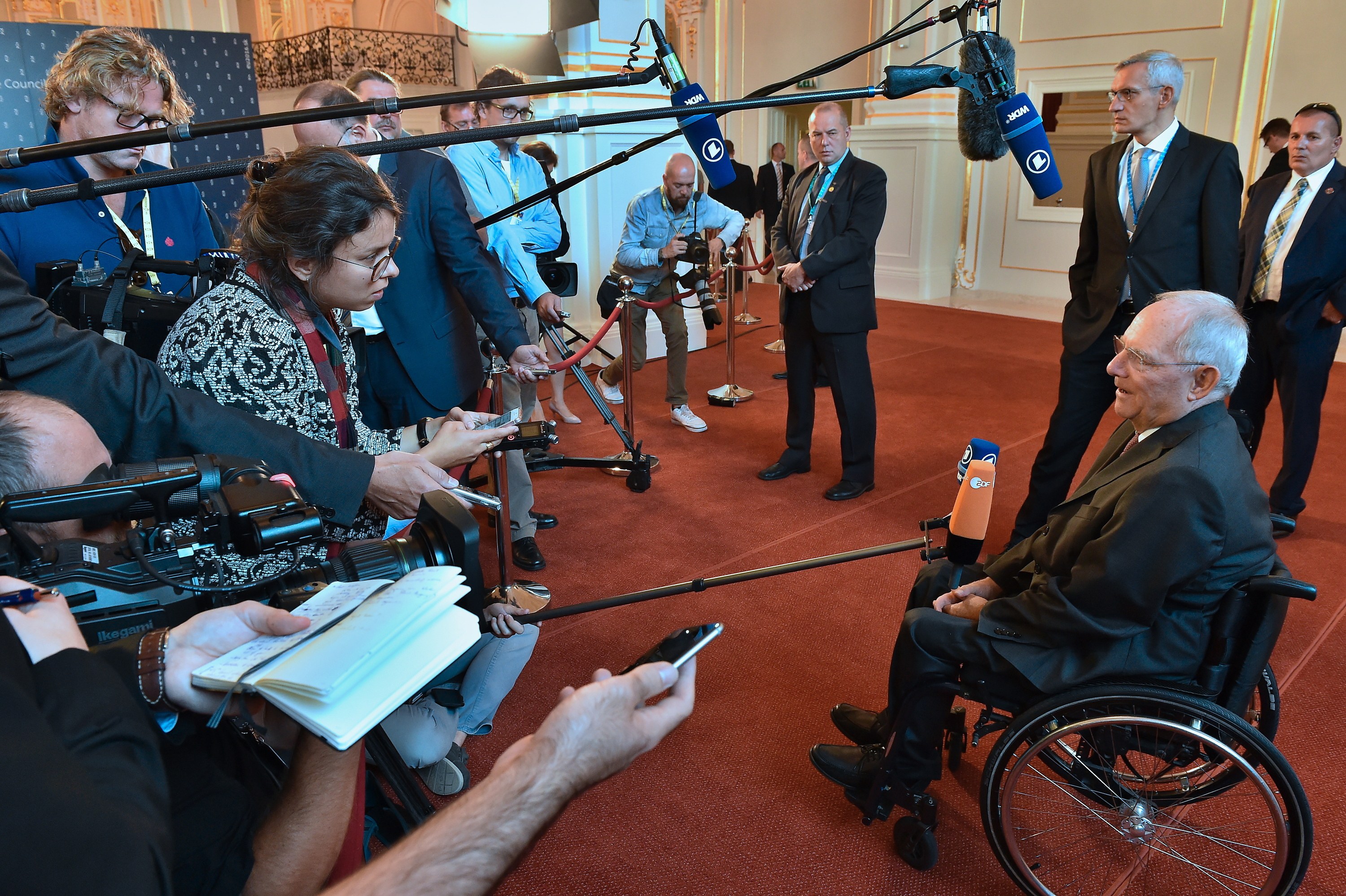 Ein Mann im Rollstuhl in Anzug und Krawatte ist von Reportern mit Kameras und Mikrofonen umgeben, mit einem Banner und einer Wand im Hintergrund.