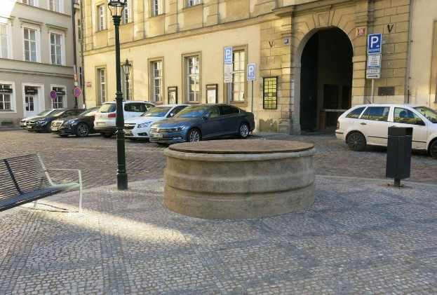 Ein Stadtplatz mit einer zentralen Bank, einem Wasserbrunnen, Laternenmasten und umliegenden Gebäuden mit Fenstern und Schildern.