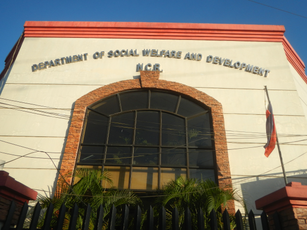 Das Gebäude des Departments für Soziales und Entwicklung mit Text an der Wand, einem Fahnenmast mit einer flatternden Flagge, einem Metallzaun, Pflanzen, Bäumen, Drähten und einem bewölkten Himmel.