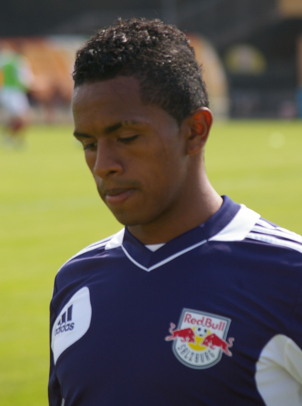 Ein junger Mann in einem blauen T-Shirt steht auf einem Fußballfeld, als Flügelspieler mit kurzen dunklen Haaren und entschlossener Miene vor einem unscharfen Hintergrund.