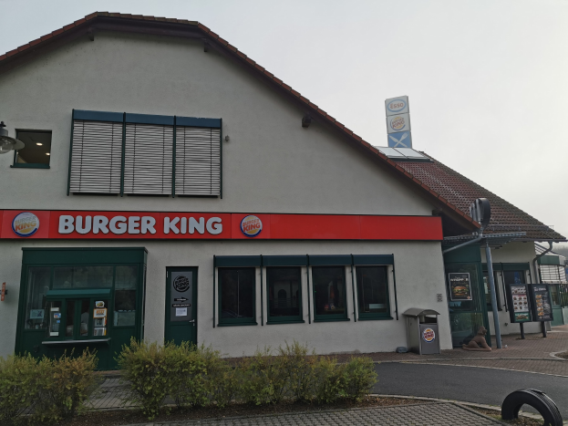 Ein Burger King Restaurant auf einer Stadtstraße mit umliegenden Gebäuden, Straßenmöbeln, Fahrzeugen, Grünflächen und einem klaren Himmel.