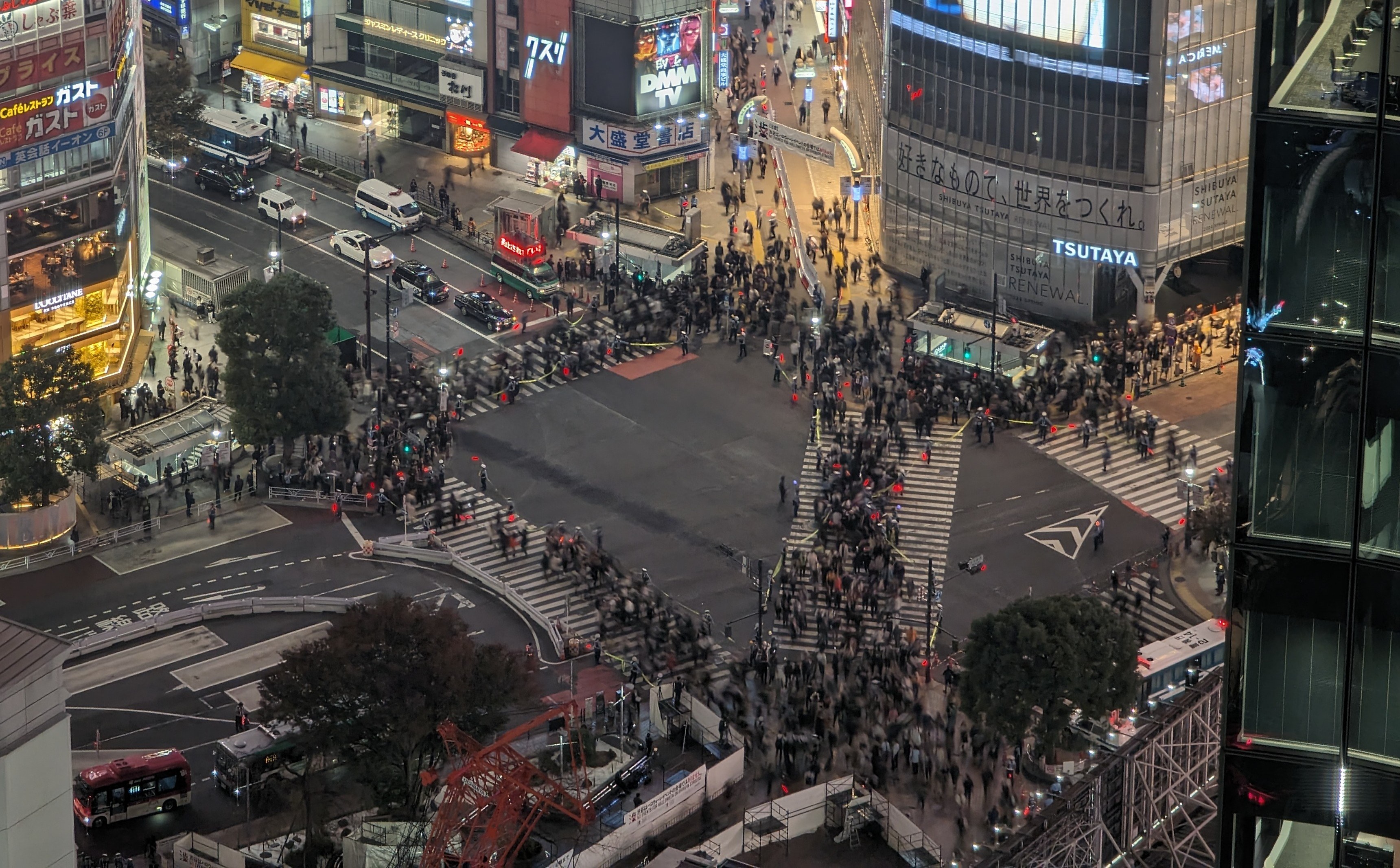 Eine nächtliche Stadtstraße mit starkem Verkehr, Passanten und einem prominenten Wolkenkratzer in der Mitte, umgeben von Gebäuden, Straßenschildern und Bäumen.
