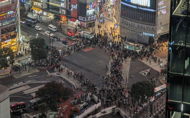 Eine nächtliche Stadtstraße mit starkem Verkehr, Passanten und einem prominenten Wolkenkratzer in der Mitte, umgeben von Gebäuden, Straßenschildern und Bäumen.