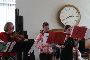 Drei Frauen auf der Bühne mit Geigen stehend und spielend, mit Notenpulten und Noten davor, während ein Zuschauer im Vordergrund sitzt; eine Uhr ist an der weißen Rückwand angebracht, und ein weißer Vorhang ist links sichtbar.