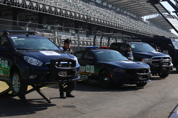 Eine Gruppe von Polizeiwagen vor einer Rennstrecke geparkt, uniformierte Personen auf der Straße stehen, im Hintergrund ein Stadion und Maschendrahtzaun.