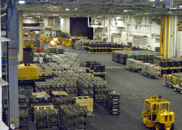 Lagerhalle mit Trucks, Containern, Menschen, Beleuchtungsgeräten, Metallstangen und einem sichtbaren Rad.