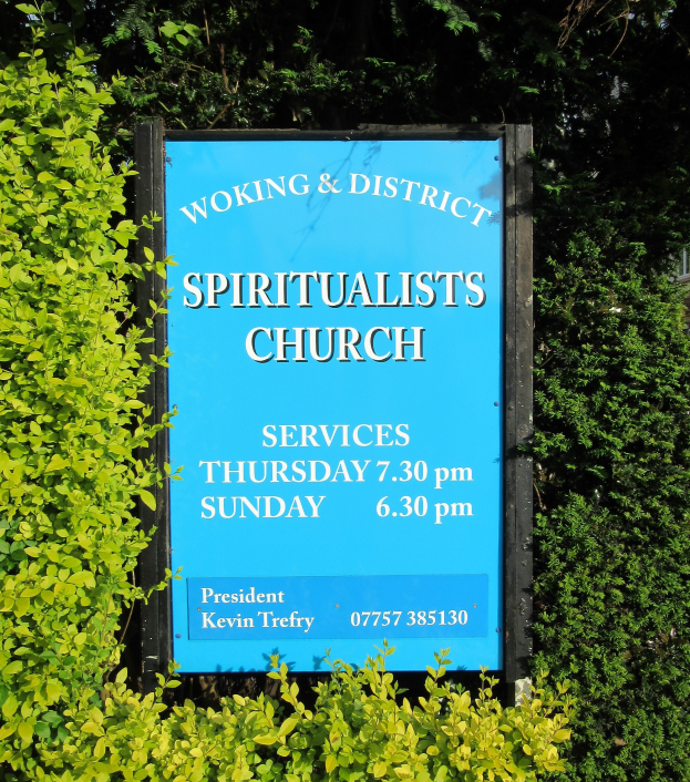 Außenansicht eines Schildes mit der Aufschrift "Woking & District Spiritualists Church" in einer Gartenumgebung mit Grünpflanzen und einem Gebäude im Hintergrund.