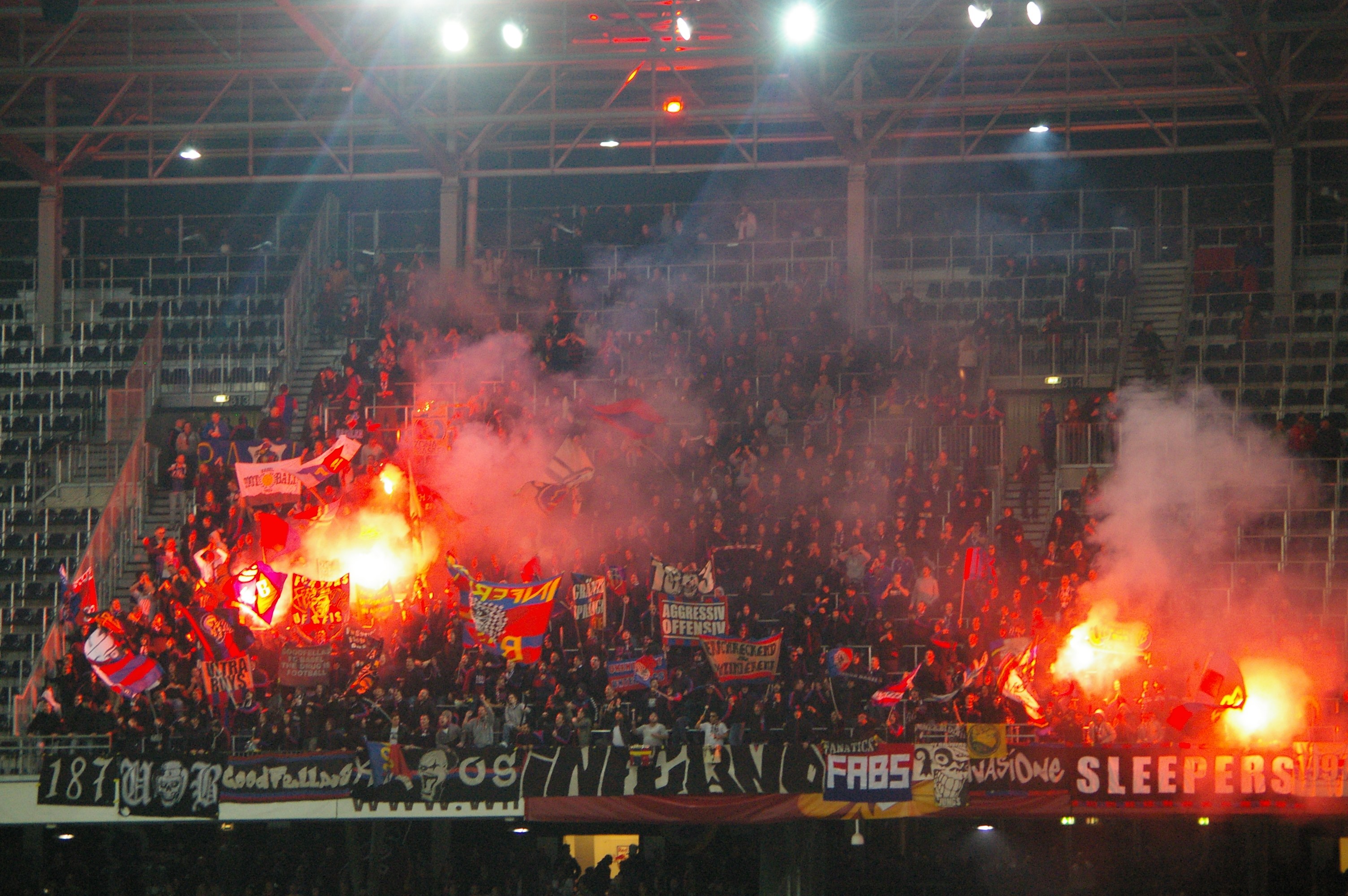 Eine große Menge Menschen in einem Stadion hält Fahnen und Banner, mit Leuchtraketen und Rauch, während Banner mit Text, Metallrahmen, Stangen und Deckenlampen sichtbar sind.