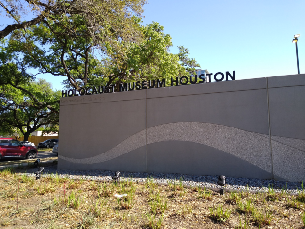 Außenansicht des Holocaust-Museums Houston mit einer Gedenkwand, Steinen, Grünflächen, Bäumen, Fahrzeugen, einer Straßenlaterne und einem bewölkten Himmel.
