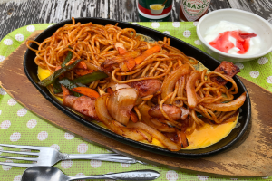 Ein Teller Nudeln mit Fleisch und Gemüse auf einem hölzernen Schneidbrett, mit einer Gabel, einem Löffel und einer Schüssel Sauce, neben Flaschen auf dem Tisch.