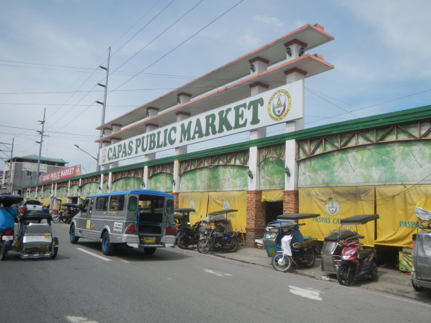 Lebendige Stadtstraße mit Autos, Motorrädern und Rikschas vor dem Gebäude "Capas Public Market", Strommasten, Laternen, Gebäuden und bewölktem Himmel im Hintergrund.