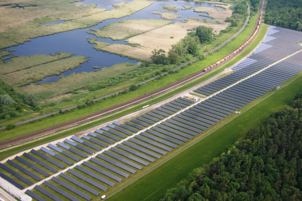 Eine Luftaufnahme eines Solarparks mit zahlreichen Solarpanelen in einem Feld, umgeben von Bäumen, Gras und Wasser, mit einem Zug, der auf einem nahen Bahngeleis fährt.