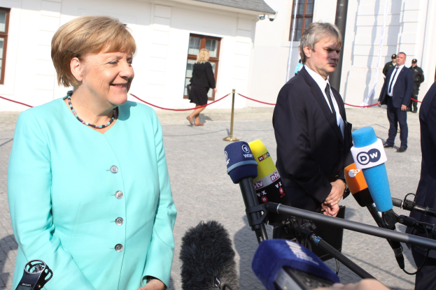 Bundeskanzlerin Angela Merkel spricht vor einem Gebäude mit Fenstern zu Reportern, umgeben von Menschen und Medienpersonal mit Mikrofonen und Ständern, eine Absperrung aus Seil ist sichtbar, bewölkter Himmel.