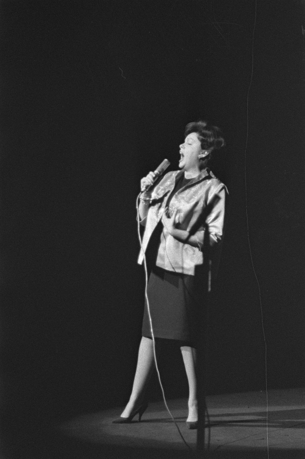 Eine Frau in einem schwarzen und weißen Outfit aus den 1950er-Jahren singt fröhlich auf einer von einem Scheinwerfer beleuchteten Bühne in ein Mikrofon, ihre Haare sind im klassischen 1950er-Stil frisiert.