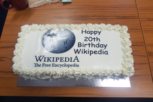 Ein Geburtstagskuchen mit der Aufschrift "Happy 20th Birthday Wikipedia" auf einem Tisch mit einer Tasse, einer Hülle und anderen Gegenständen.