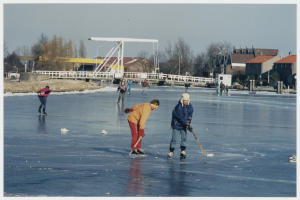 Eine Gruppe von Menschen, die Eisstockschießen auf einem zugefrorenen See betreiben, umgeben von Bäumen, Gebäuden, Pfählen und einer Brücke, unter einem von Straßenlaternen beleuchteten Himmel.