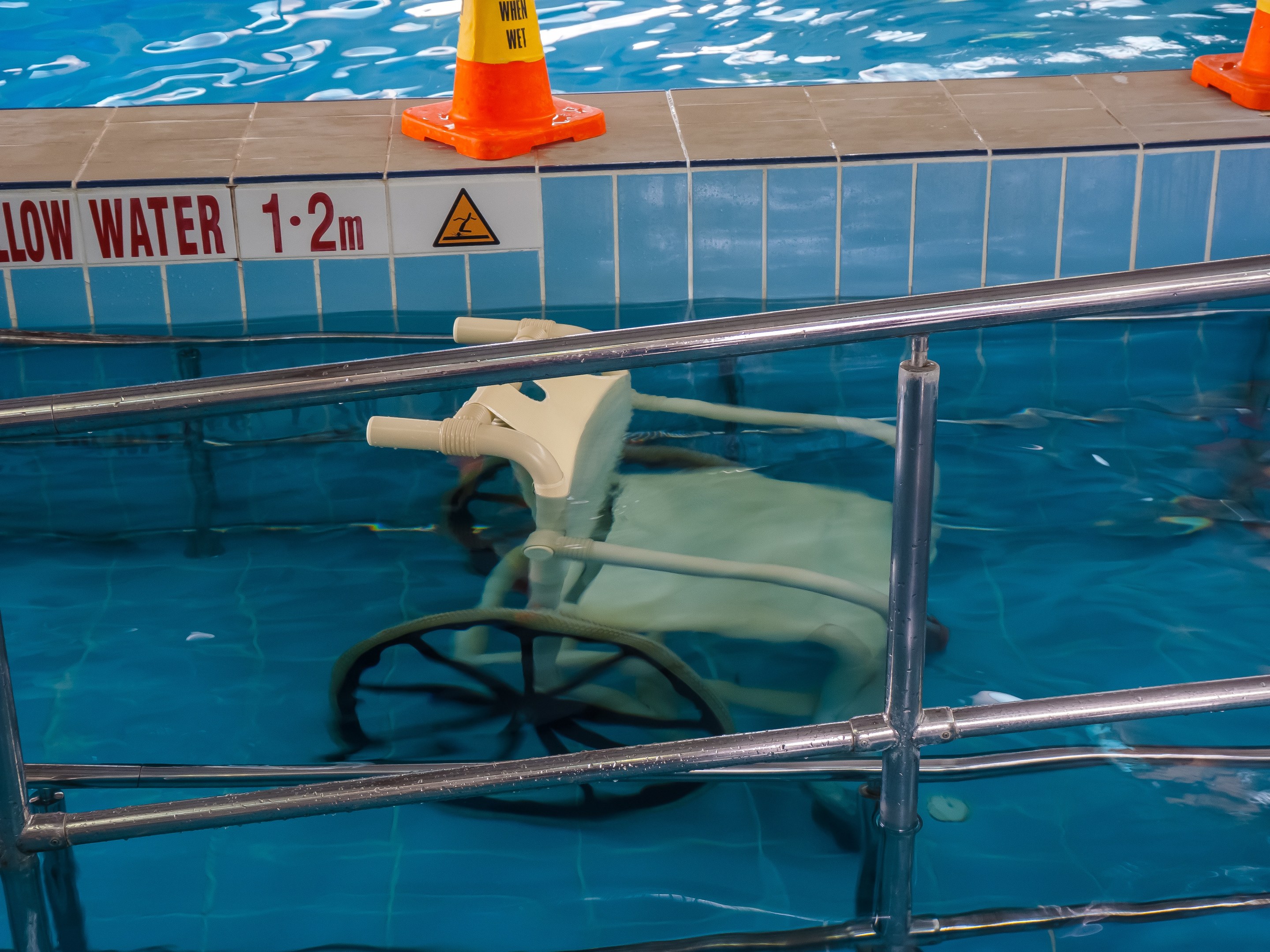 Schwimmbad mit einem Stuhl in der Mitte, umgeben von Sicherheitskegeln und einem Geländer, sowie einem Schild an der Wand, das verfügbare Schwimmausrüstung anzeigt.