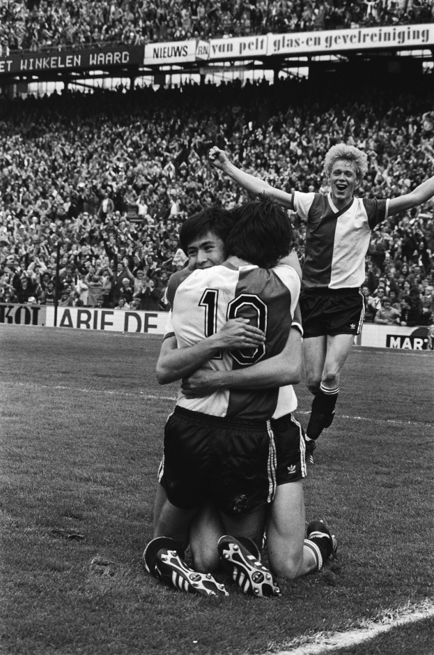 Drei Fuballspieler umarmen sich auf dem Boden nach einem Tor, mit jubelnder Menge und schwenkenden Fahnen im Hintergrund, in einer schwarz-weißen Stadionatmosphäre.
