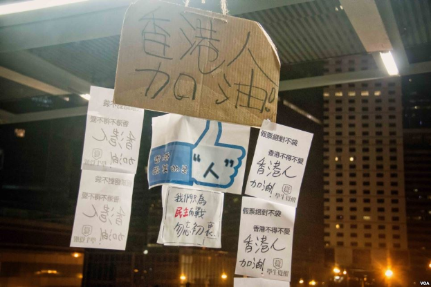 Eine Gruppe von Menschen steht vor einem beleuchteten Gebäude in der Nacht, mit Protestschildern an der Decke.