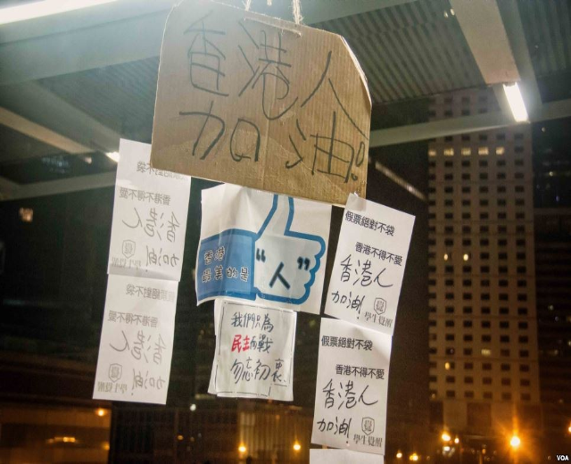 Eine Gruppe von Menschen steht vor einem beleuchteten Gebäude in der Nacht, mit Protestschildern an der Decke.